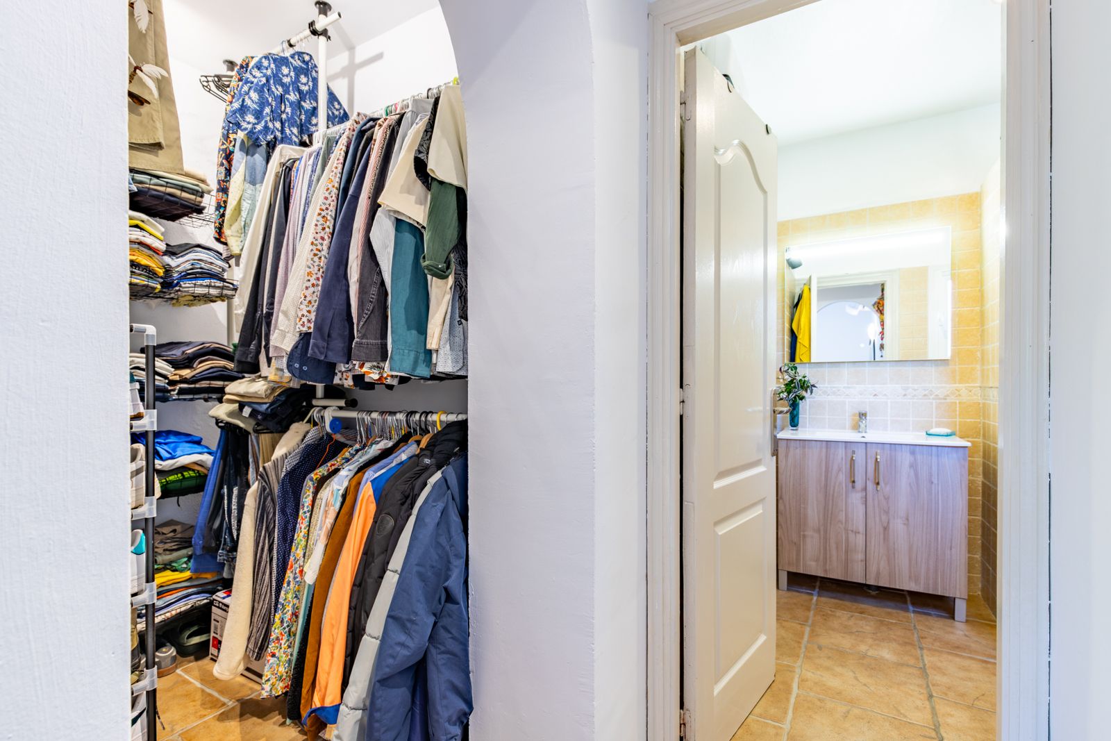 Dressing ouvert avec étagères et penderies, offrant un accès direct à une salle de bain avec meuble en bois et miroir rétroéclairé.