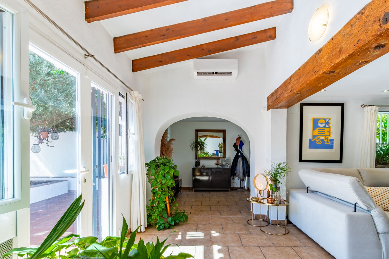 Espace de vie lumineux avec poutres apparentes, sol en terre cuite et accès terrasse. Une arche relie l'entrée sous un plafond incliné équipé de la climatisation.