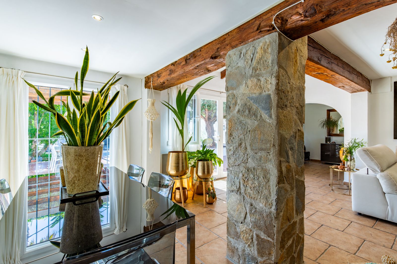 Espace repas avec poutres en bois et pilier en pierre naturelle. Sols en terre cuite, table en verre et grandes ouvertures apportant une luminosité naturelle.