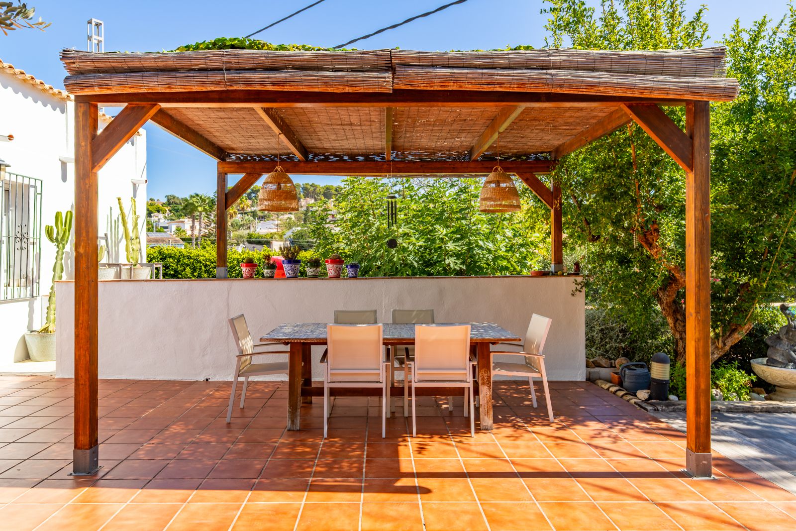 Espace repas extérieur abrité par une pergola en bois avec toiture en canisse, suspensions en osier et carrelage en terre cuite.