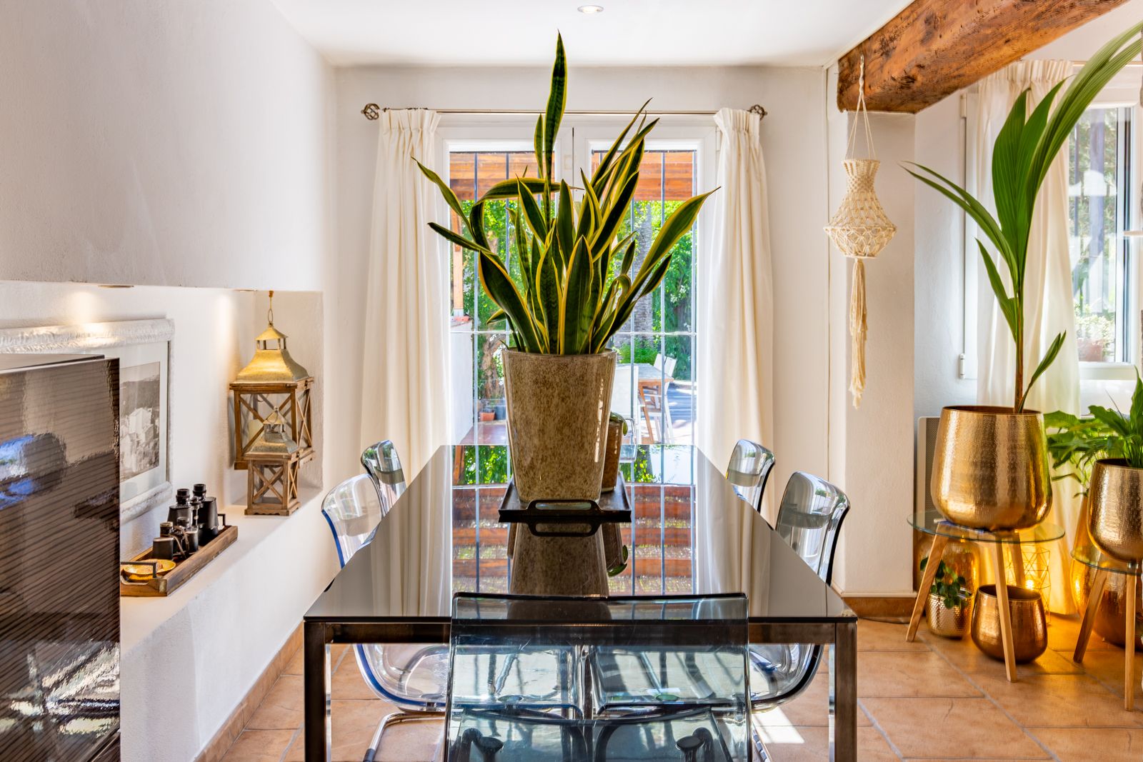 Salle à manger lumineuse avec table en verre, chaises transparentes et poutres en bois. Les portes-fenêtres offrent une vue et un accès direct à l'extérieur.
