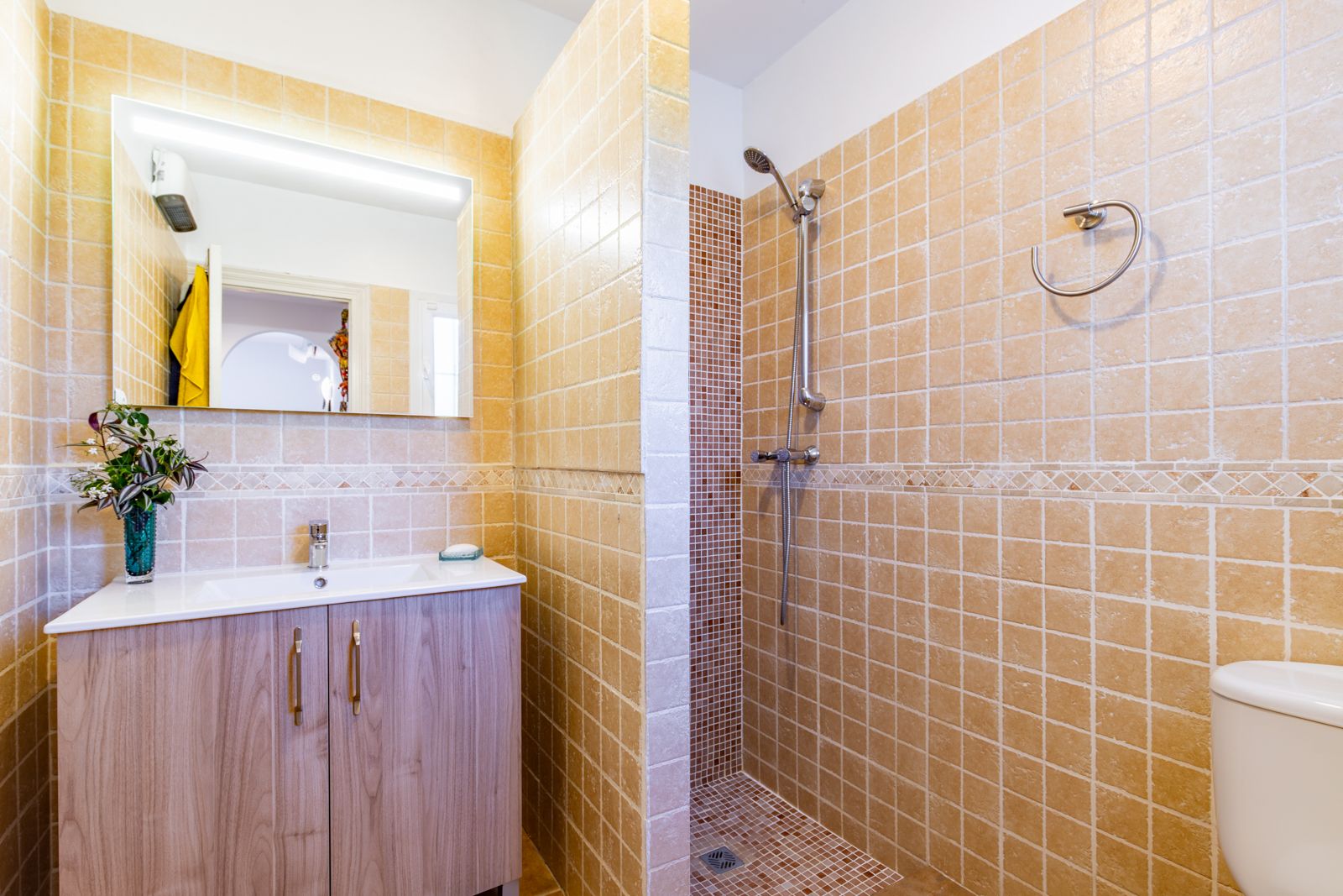 Salle de bain avec douche à l'italienne en mosaïque et carrelage aux tons ocre. Comprend un meuble vasque à finition bois et un miroir avec éclairage intégré.