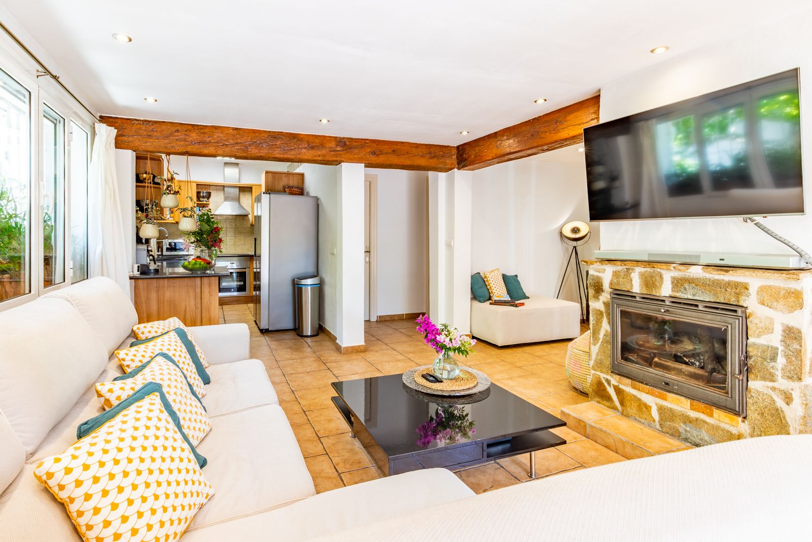 Salon lumineux avec poutres en bois apparentes, cheminée en pierre naturelle et sol en terre cuite, ouvert sur une cuisine fonctionnelle.