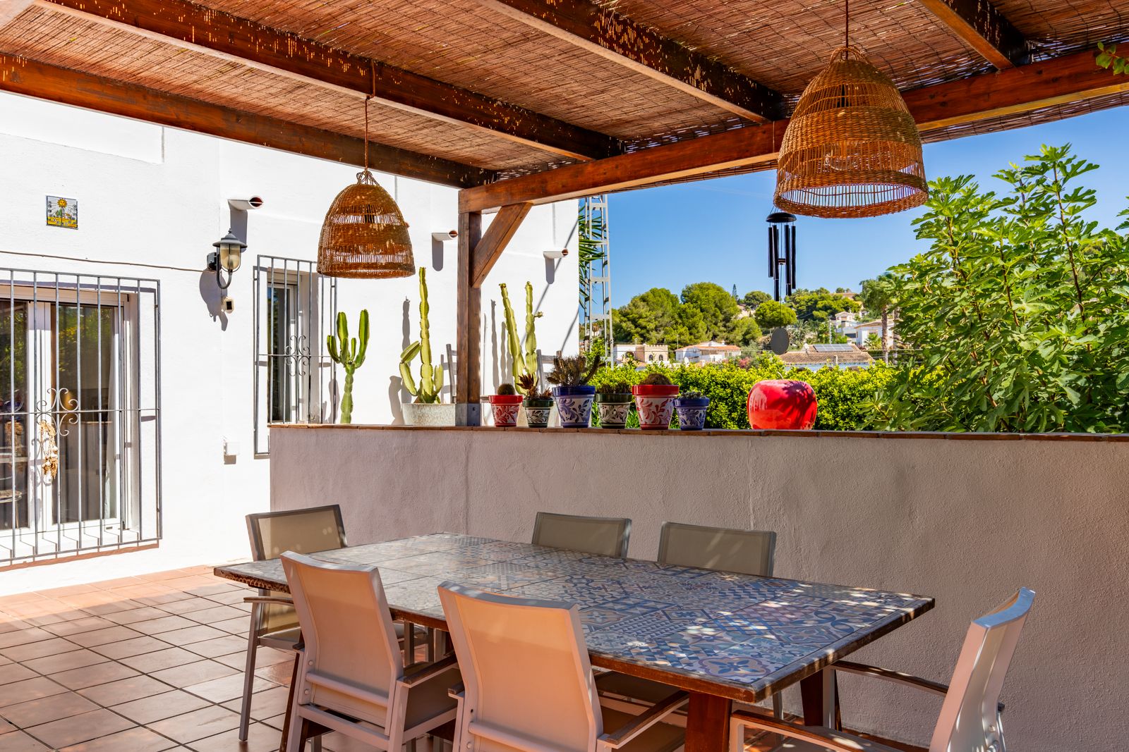 Terrasse couverte avec pergola en bois et canisse, table à manger en céramique décorée et suspensions en osier offrant une vue dégagée sur la végétation.