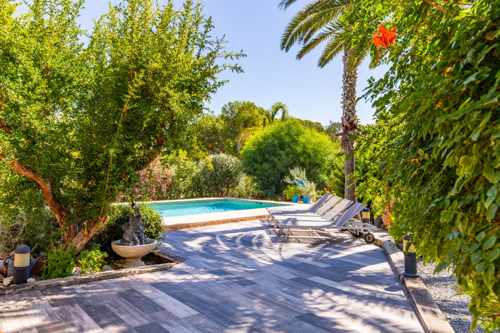 Terrasse en grès cérame aspect bois avec chaises longues, bordée d'une végétation méditerranéenne luxuriante et d'une piscine privée ensoleillée.