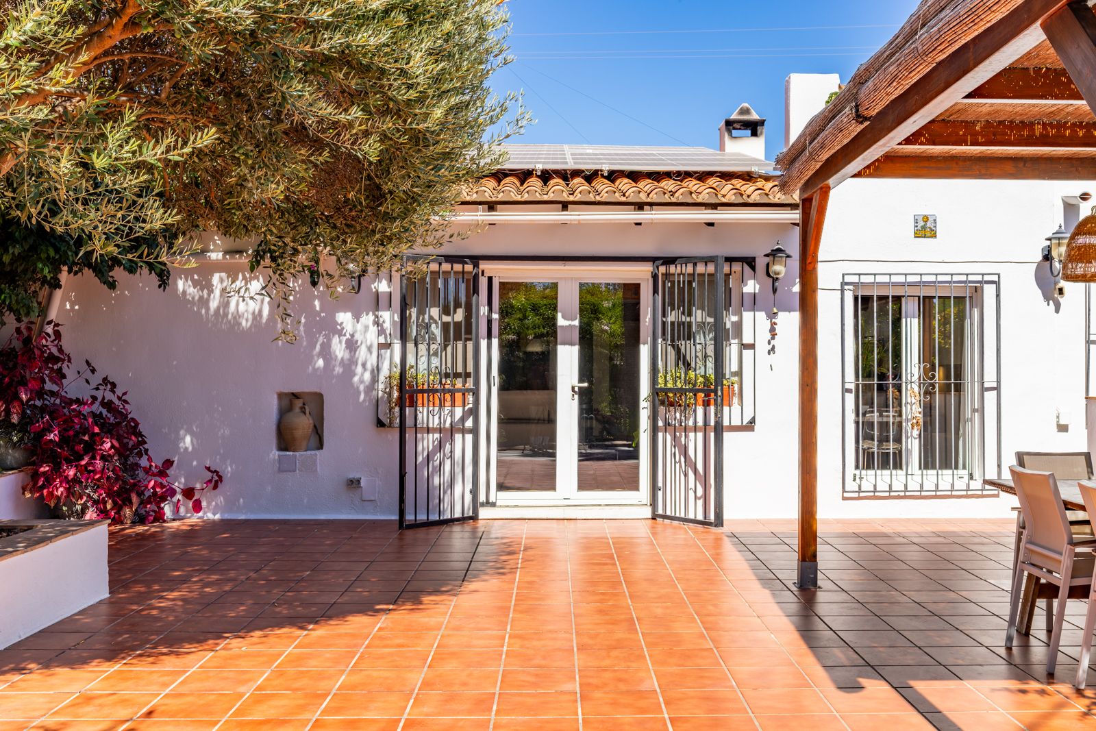 Terrasse extérieure avec carrelage en terre cuite, pergola en bois et panneaux solaires. La façade blanche intègre des ferronneries noires et un olivier apportant de l'ombre.
