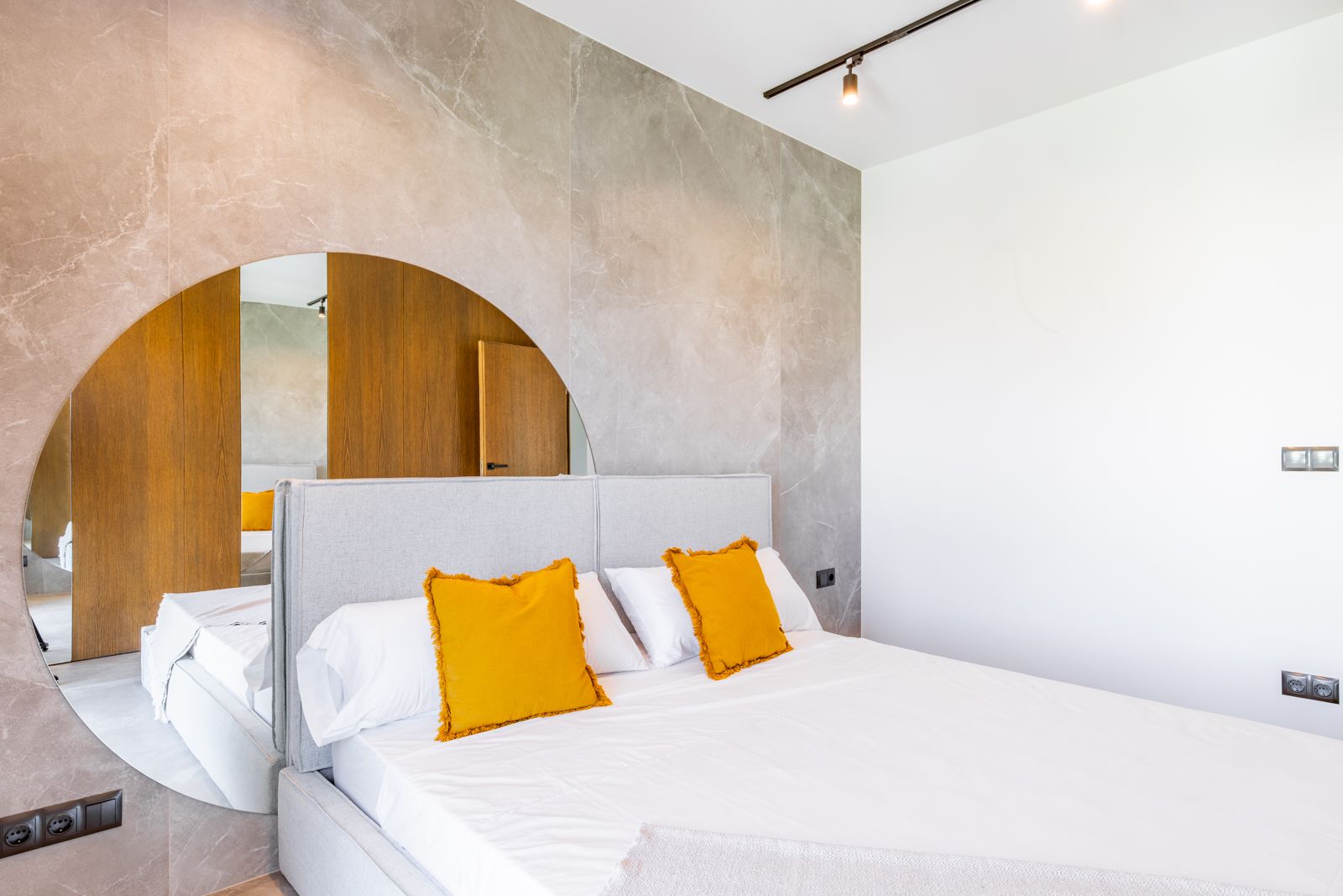 Chambre avec mur en grès cérame et miroir circulaire. Elle dispose d'une tête de lit en tissu, de finitions en chêne naturel et d'un éclairage sur rail moderne.