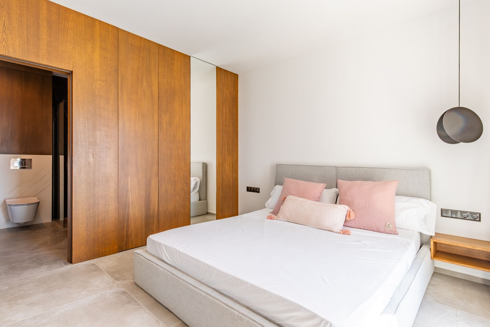Chambre avec placards sur mesure en chêne naturel, sol en grès cérame et salle de bain attenante équipée de sanitaires suspendus modernes.