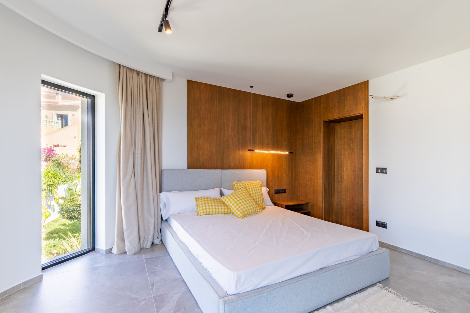 Chambre avec revêtement mural en chêne naturel, éclairage suspendu moderne et sol en grès cérame. La grande baie vitrée s'ouvre sur un jardin méditerranéen soigné.
