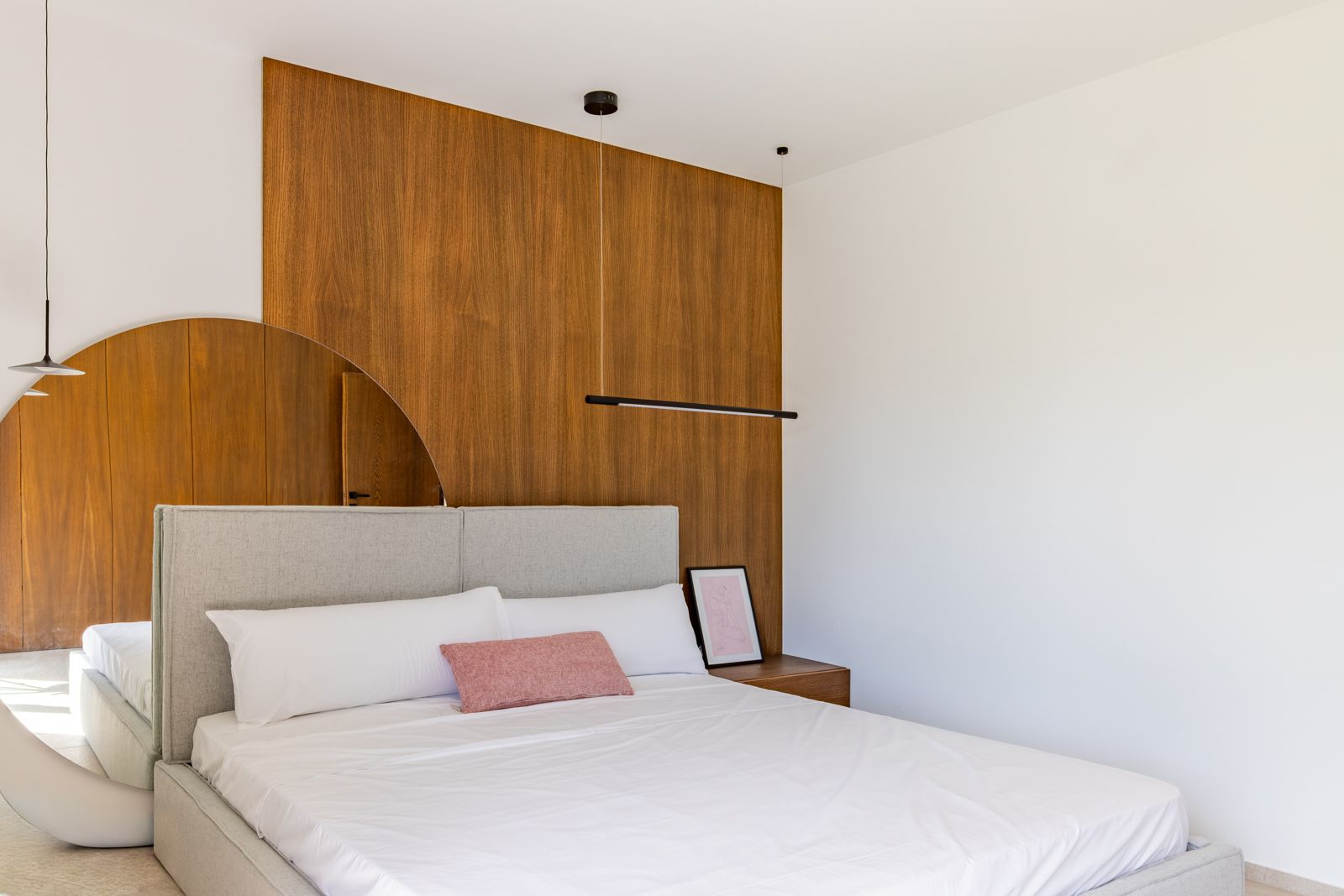 Chambre moderne avec revêtement mural en chêne naturel, tête de lit en textile gris et grand miroir circulaire. Éclairage suspendu minimaliste et table de chevet intégrée.
