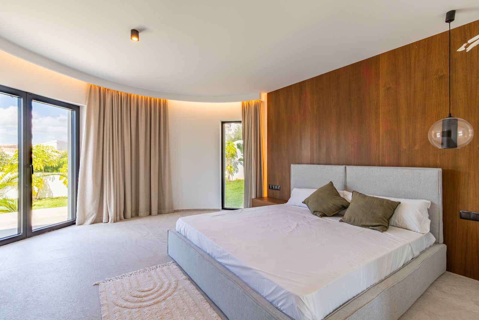 Chambre principale avec revêtement mural en chêne naturel, plafond incurvé à éclairage LED indirect et accès direct au jardin par de larges baies vitrées.