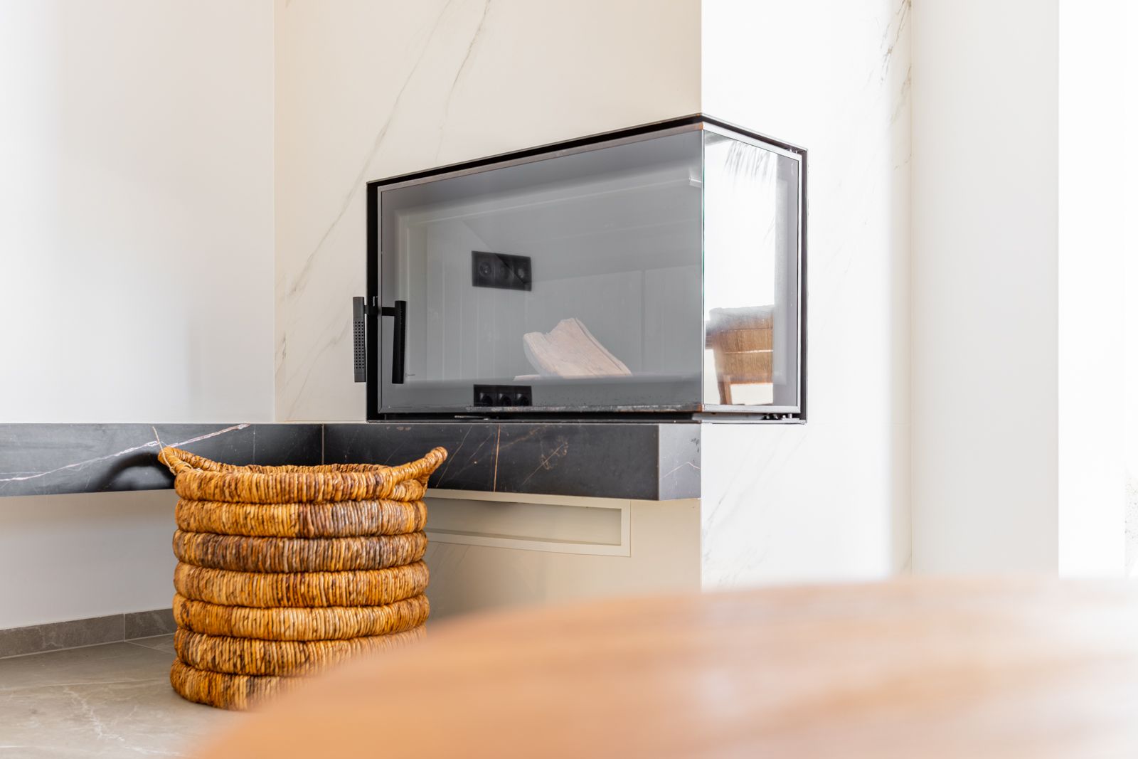 Cheminée d'angle moderne avec insert vitré, intégrée dans un revêtement en pierre frittée blanche et tablette en marbre noir, accompagnée d'un panier en osier.
