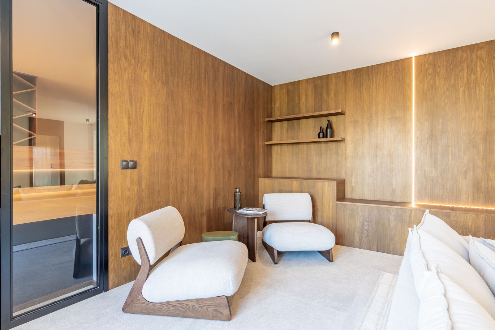 Coin salon avec revêtement mural en chêne naturel, éclairage LED intégré et sol en grès cérame. Mobilier design comprenant des fauteuils blancs élégants.