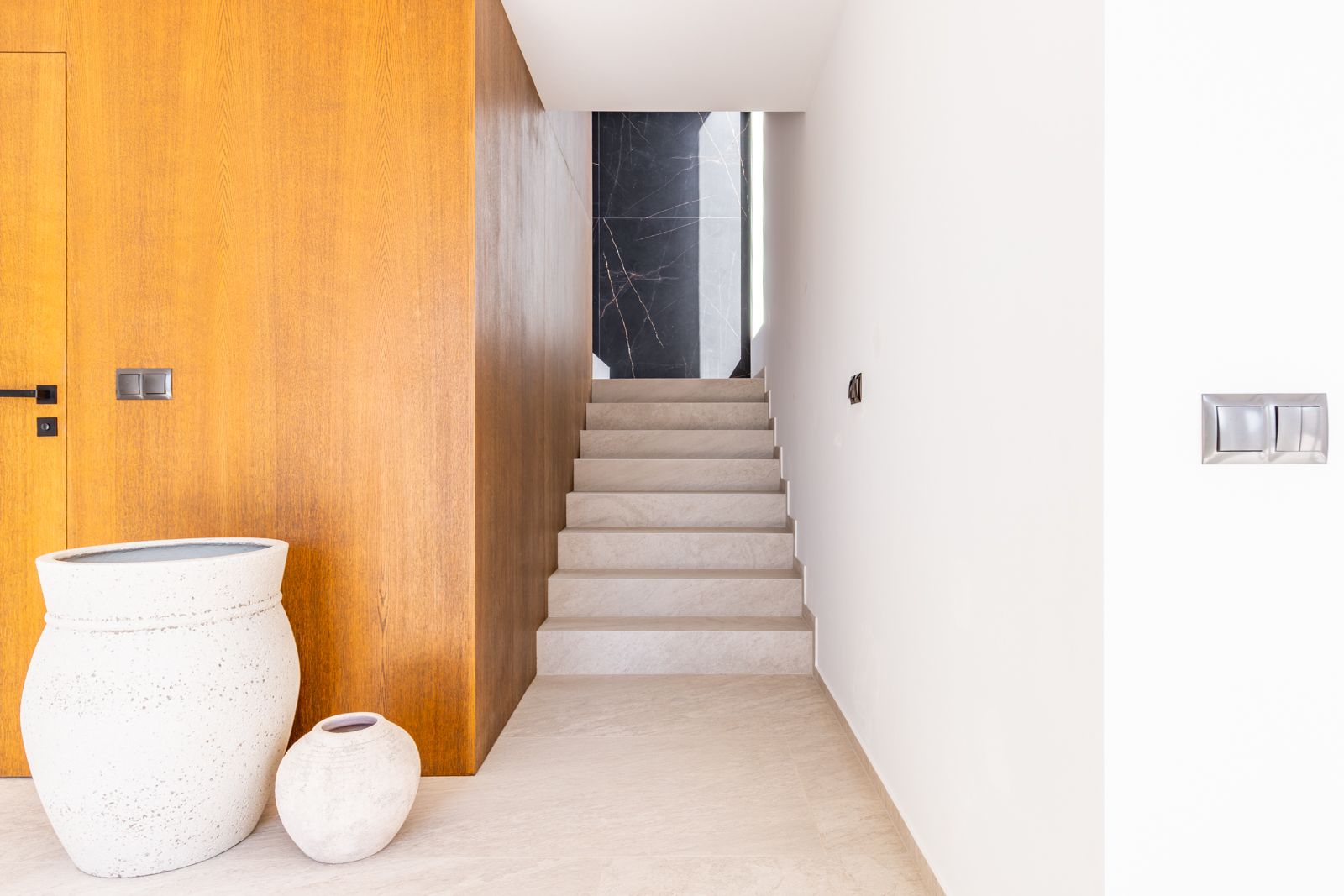Couloir avec escalier en grès cérame, mur habillé de placage en chêne naturel et paroi de fond en pierre frittée noire veinée.