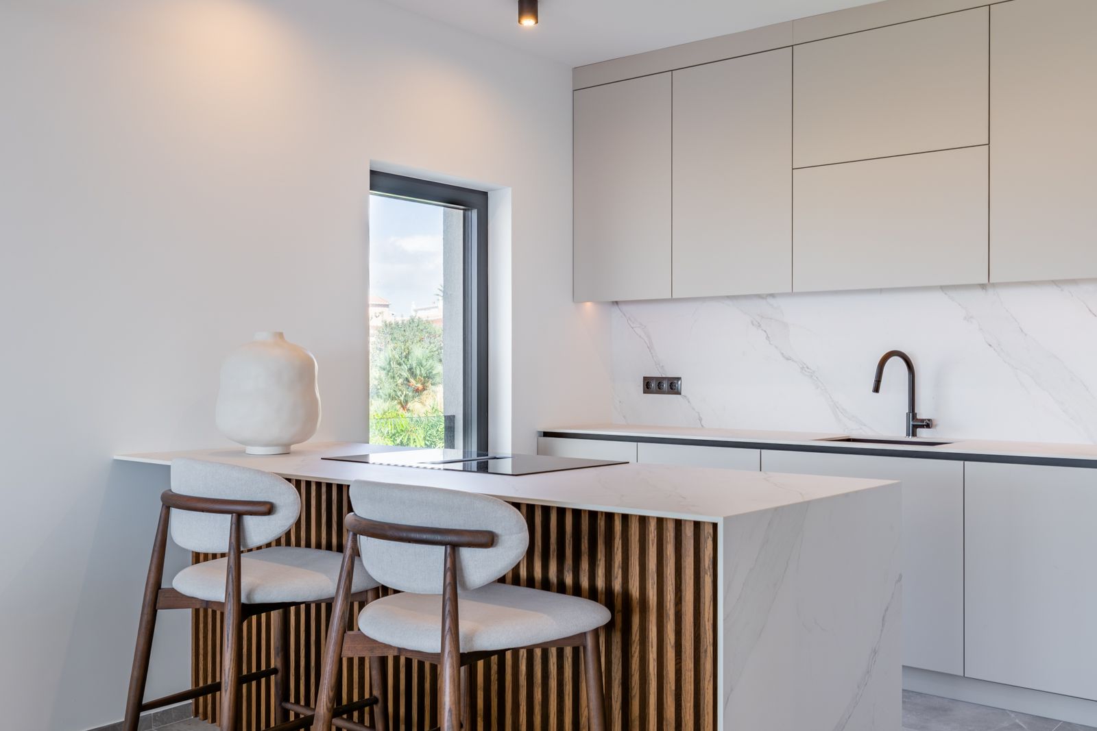 Cuisine de design avec îlot en pierre frittée effet marbre, habillage en tasseaux de bois et tabourets élégants. Équipée d'une plaque à induction et d'une robinetterie noire.