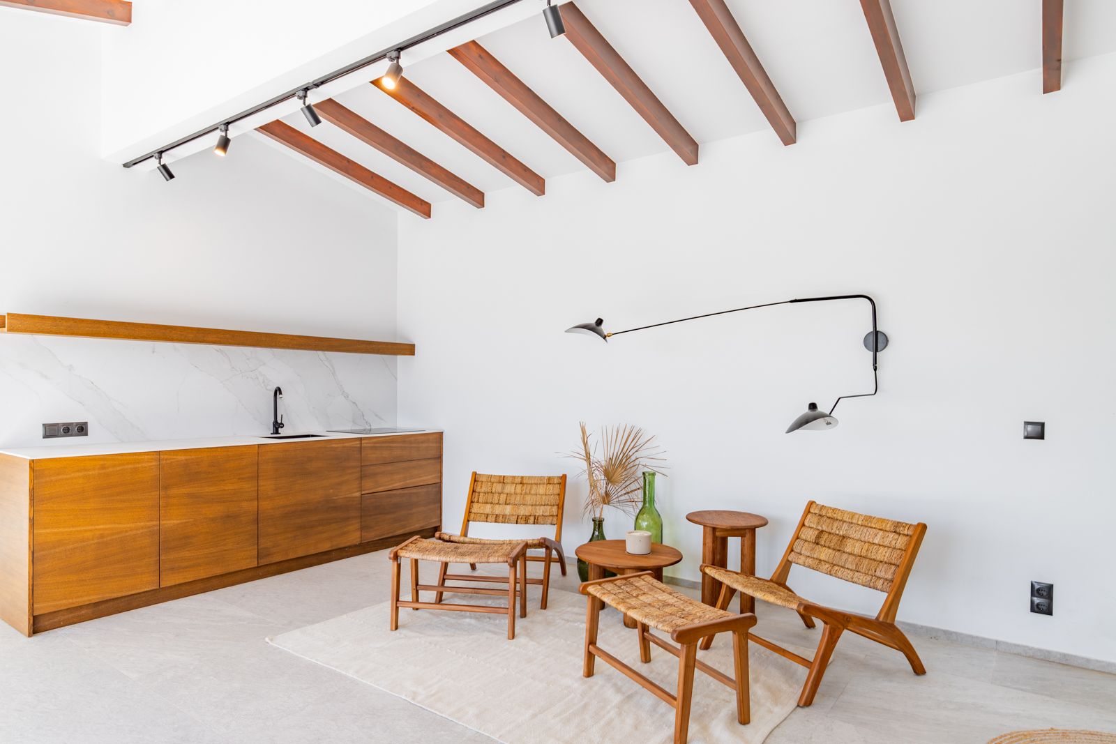 Espace de vie avec kitchenette en chêne, plan de travail en pierre frittée, poutres apparentes et fauteuils en fibres naturelles avec éclairage sur rail.
