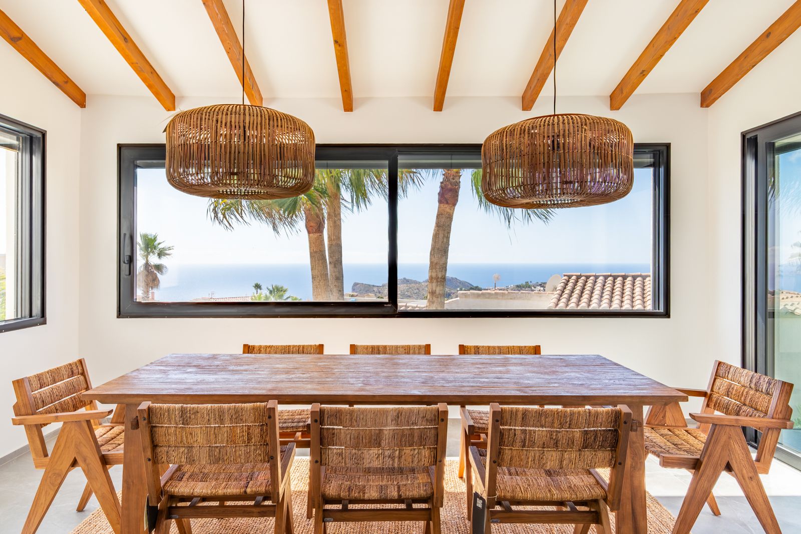 Espace repas avec poutres apparentes, table en bois massif et suspensions en osier. La large baie vitrée offre une vue dégagée sur la mer et les palmiers.