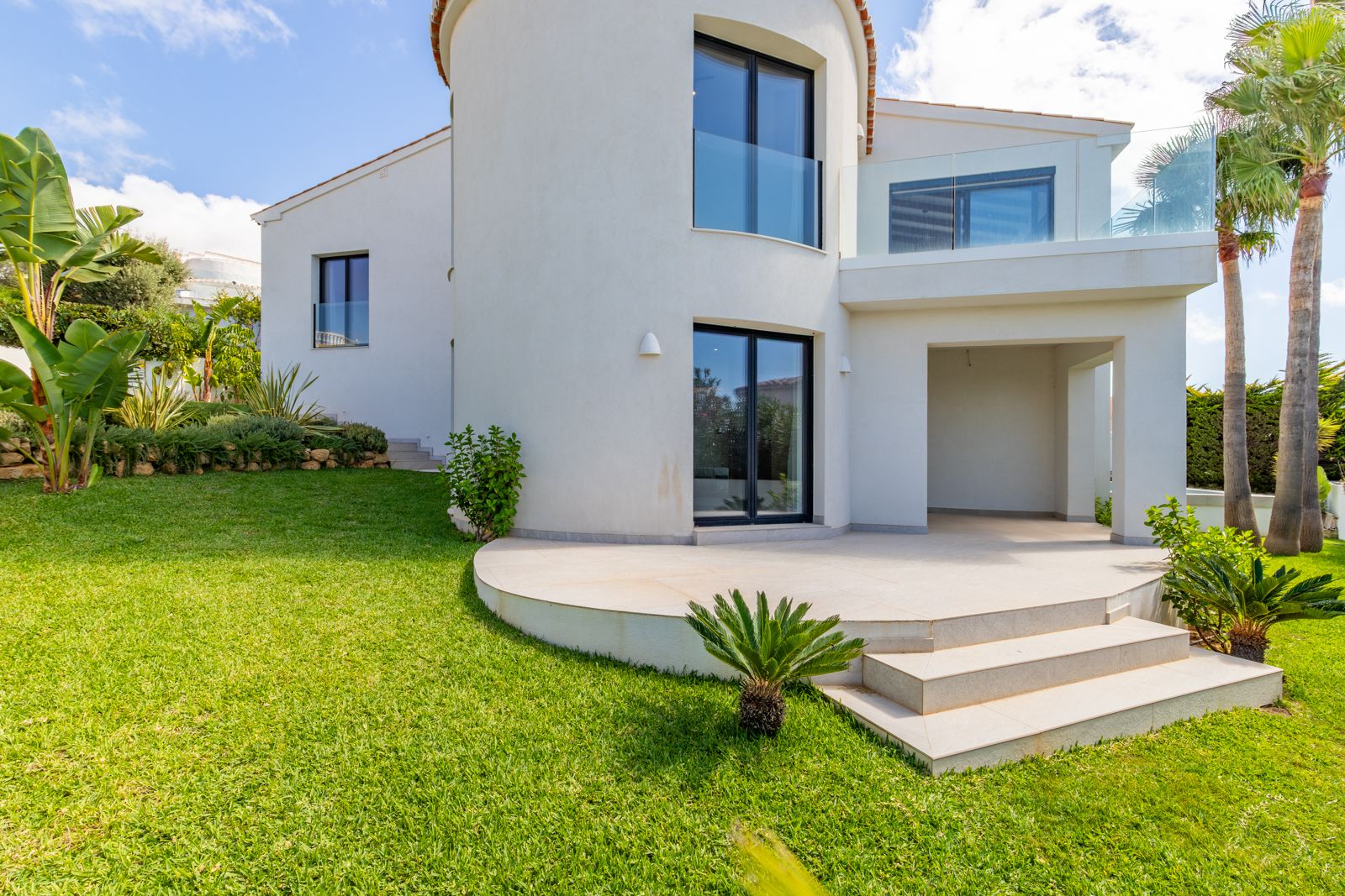 Façade arrière avec tour incurvée, menuiseries en aluminium noir et balcons à garde-corps en verre. Le jardin paysager comprend une pelouse soignée et des palmiers.