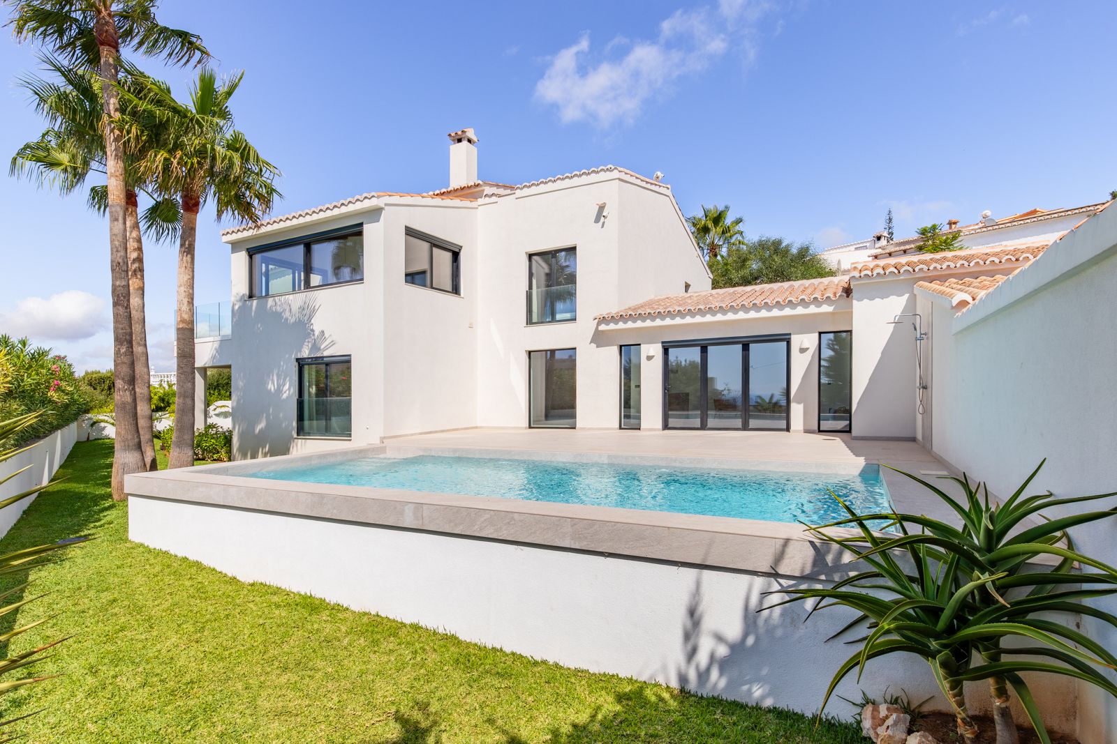 Façade arrière de la villa avec piscine au sel, terrasse en grès cérame et fenêtres à triple vitrage. Jardin paysager avec pelouse et palmiers.
