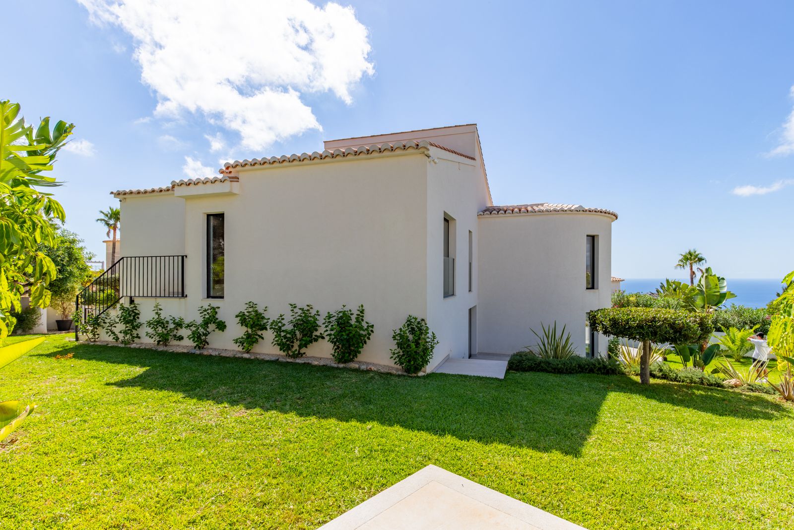 Façade blanche au design méditerranéen avec toiture en tuiles, jardin engazonné et vue sur mer, entourée d'une végétation tropicale soignée.