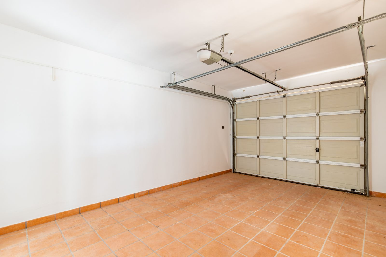 Garage fermé avec sol en grès cérame de couleur terre cuite, murs blancs lisses et porte sectionnelle motorisée avec système d'ouverture automatique au plafond.