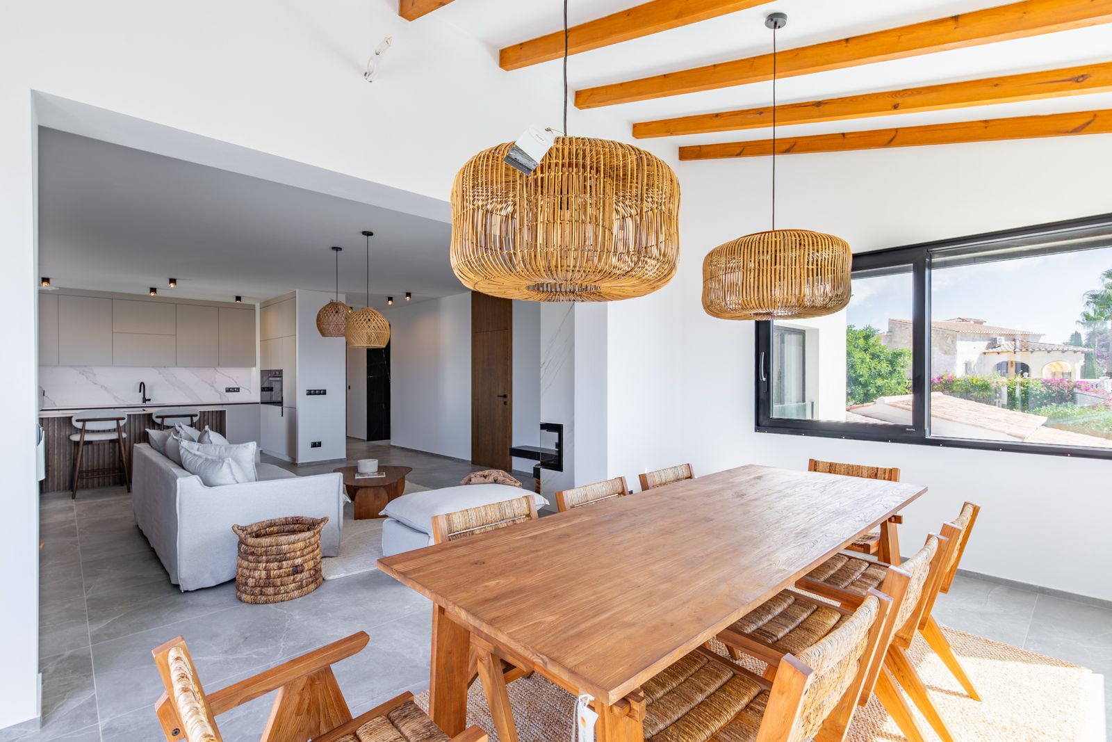 Salle à manger à aire ouverte avec table en bois massif, poutres apparentes et suspensions en rotin, intégrée au salon et à la cuisine moderne.