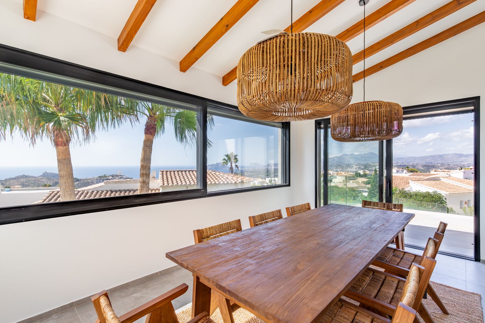 Salle à manger lumineuse avec poutres apparentes, table en bois massif et suspensions en rotin. Les larges baies vitrées offrent une vue panoramique sur la mer et les montagnes.