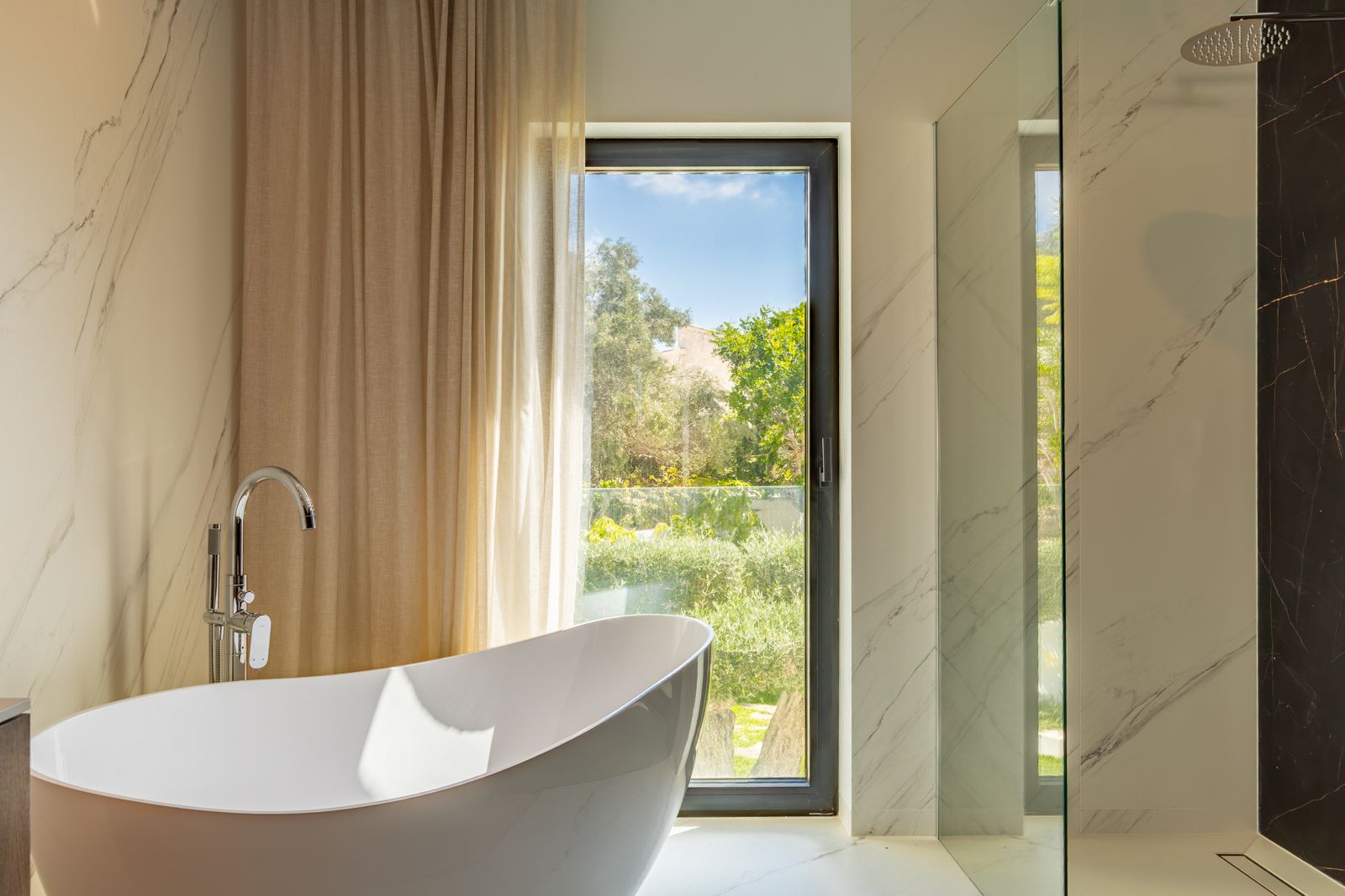 Salle de bain de design avec baignoire îlot, robinetterie sur pied et douche à l'italienne. Revêtements en grès cérame effet marbre et vue sur le jardin extérieur.