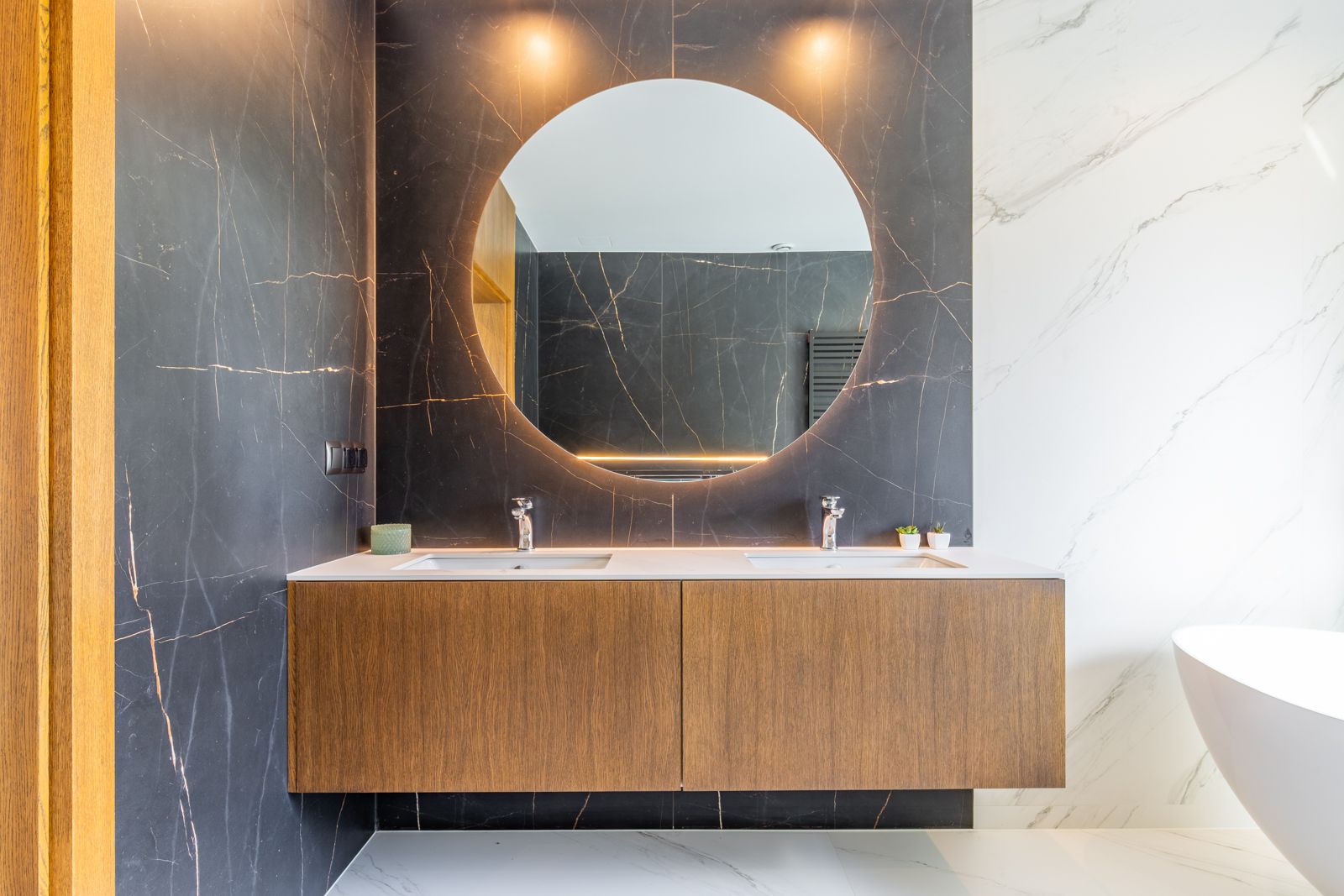 Salle de bain design avec meuble suspendu en chêne, double vasque et miroir circulaire rétroéclairé sur un mur en grès cérame foncé aux veines dorées.