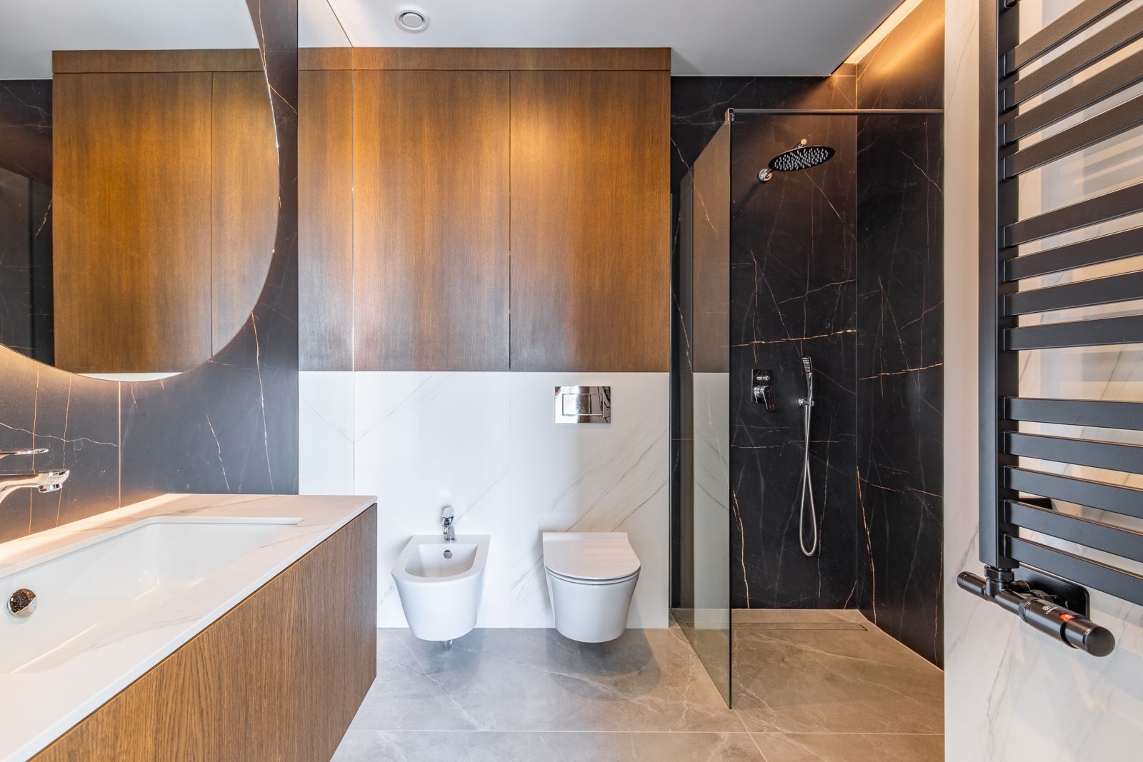 Salle de bain moderne avec revêtement en cérame foncé, douche à l'italienne, sanitaires suspendus et meubles en chêne naturel sous un grand miroir circulaire rétroéclairé.