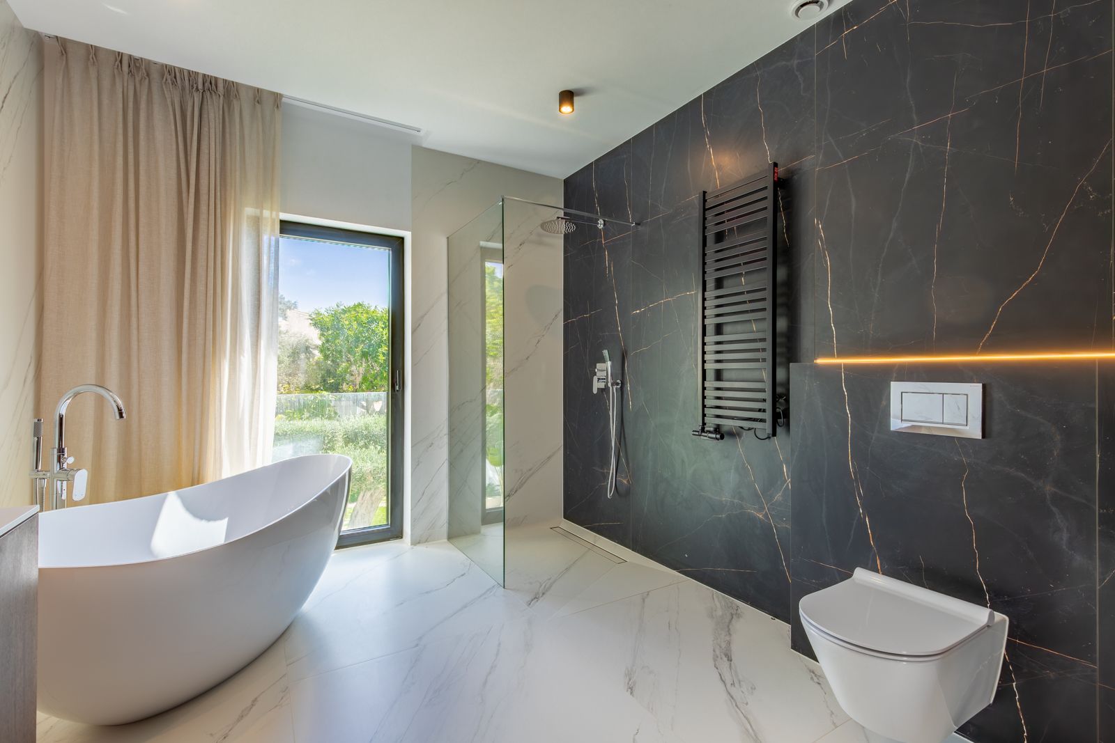 Salle de bains de design avec baignoire îlot, douche à l'italienne et grès cérame contrasté. Équipée d'un WC suspendu, d'un sèche-serviettes noir et d'une vue sur le jardin.
