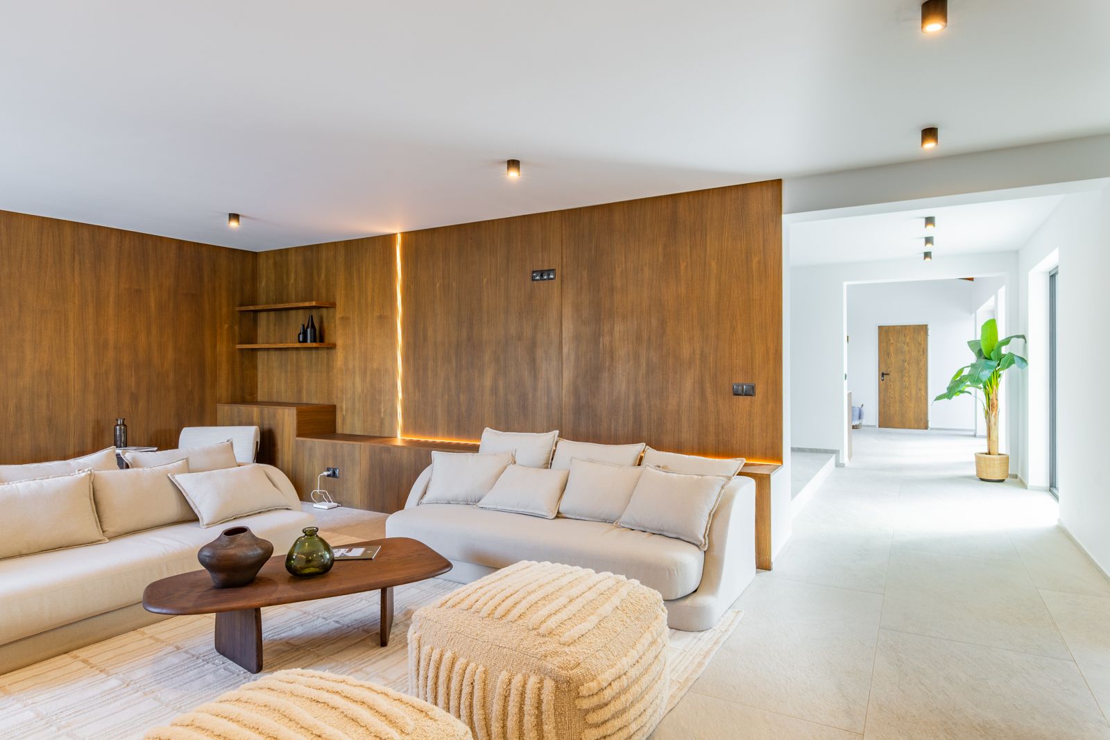 Salon avec panneaux en chêne naturel et éclairage LED intégré. Mobilier aux tons neutres sur sol en grès cérame grand format et spots de plafond cylindriques.