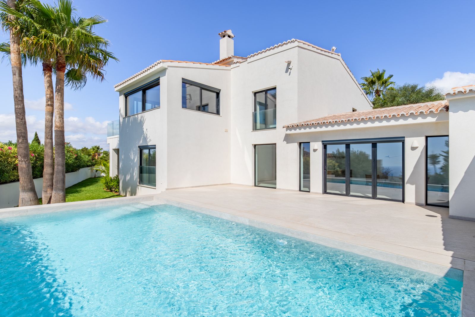 Vue extérieure avec piscine au sel et vaste terrasse en grès cérame. La façade blanche est équipée de menuiseries en aluminium et de fenêtres à triple vitrage haute performance.