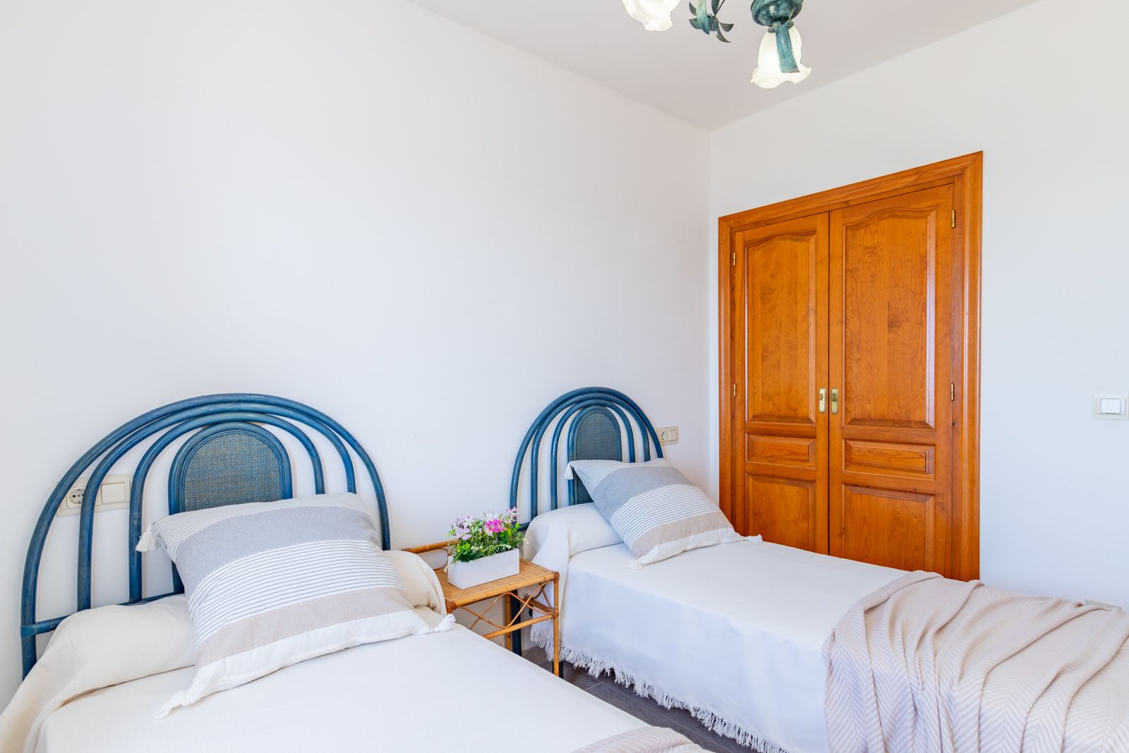 Chambre avec deux lits simples, têtes de lit en osier bleu et placard intégré en bois. Un espace lumineux aux murs blancs et au sol en carrelage traditionnel.