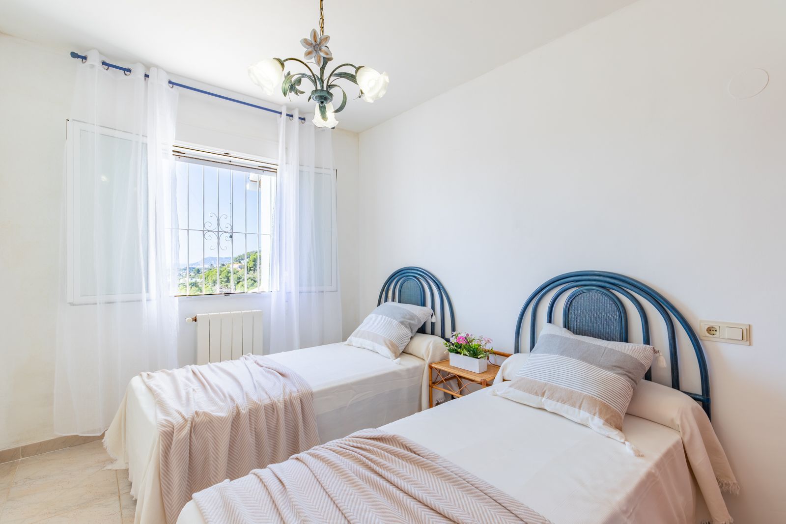 Chambre lumineuse avec deux lits simples, têtes de lit bleues arrondies, chauffage central et fenêtre ouvrant sur les collines environnantes de Benimeit.