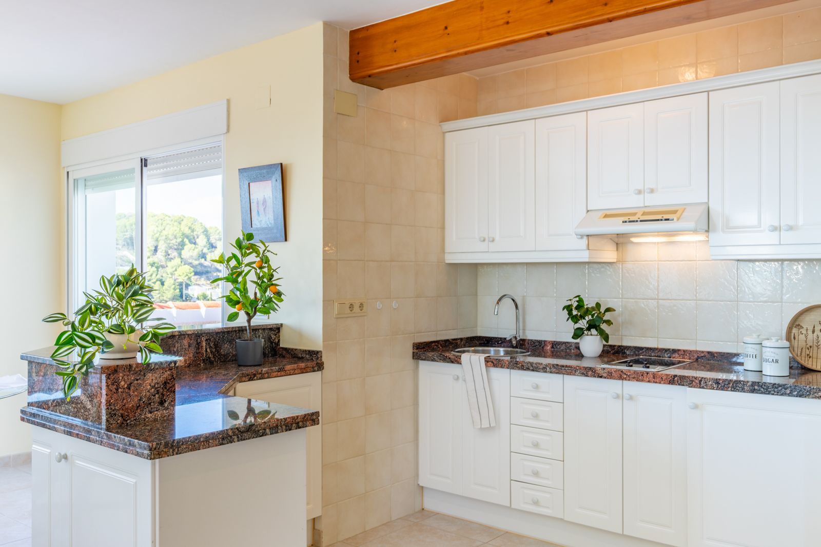 Cuisine fonctionnelle avec plans de travail en granit, meubles blancs et poutre en bois apparente. La grande fenêtre offre une luminosité naturelle et une vue sur le paysage.