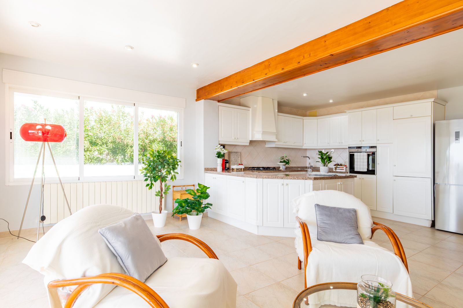 Cuisine ouverte avec meubles blancs et plan de travail en granit, intégrée dans un séjour lumineux avec poutres en bois et grandes fenêtres sur le jardin.