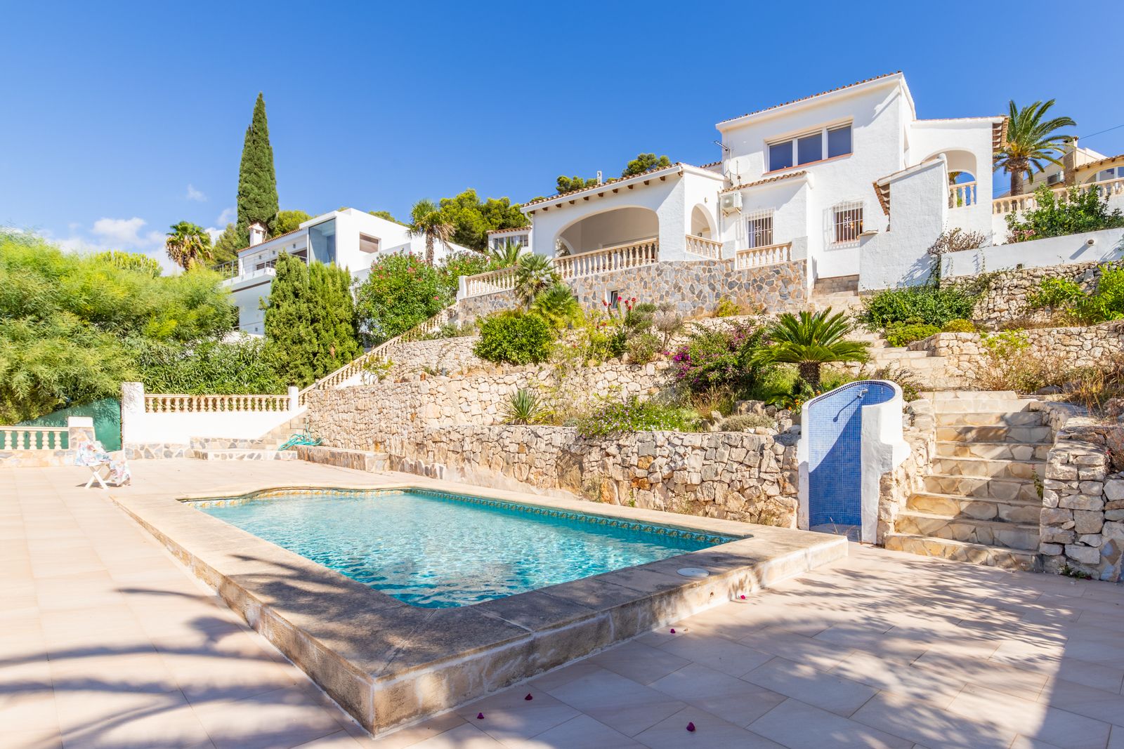 Piscine privée avec terrasse en pierre, entourée de jardins en restanques avec murs en pierre naturelle et douche extérieure sous le soleil de la Costa Blanca.