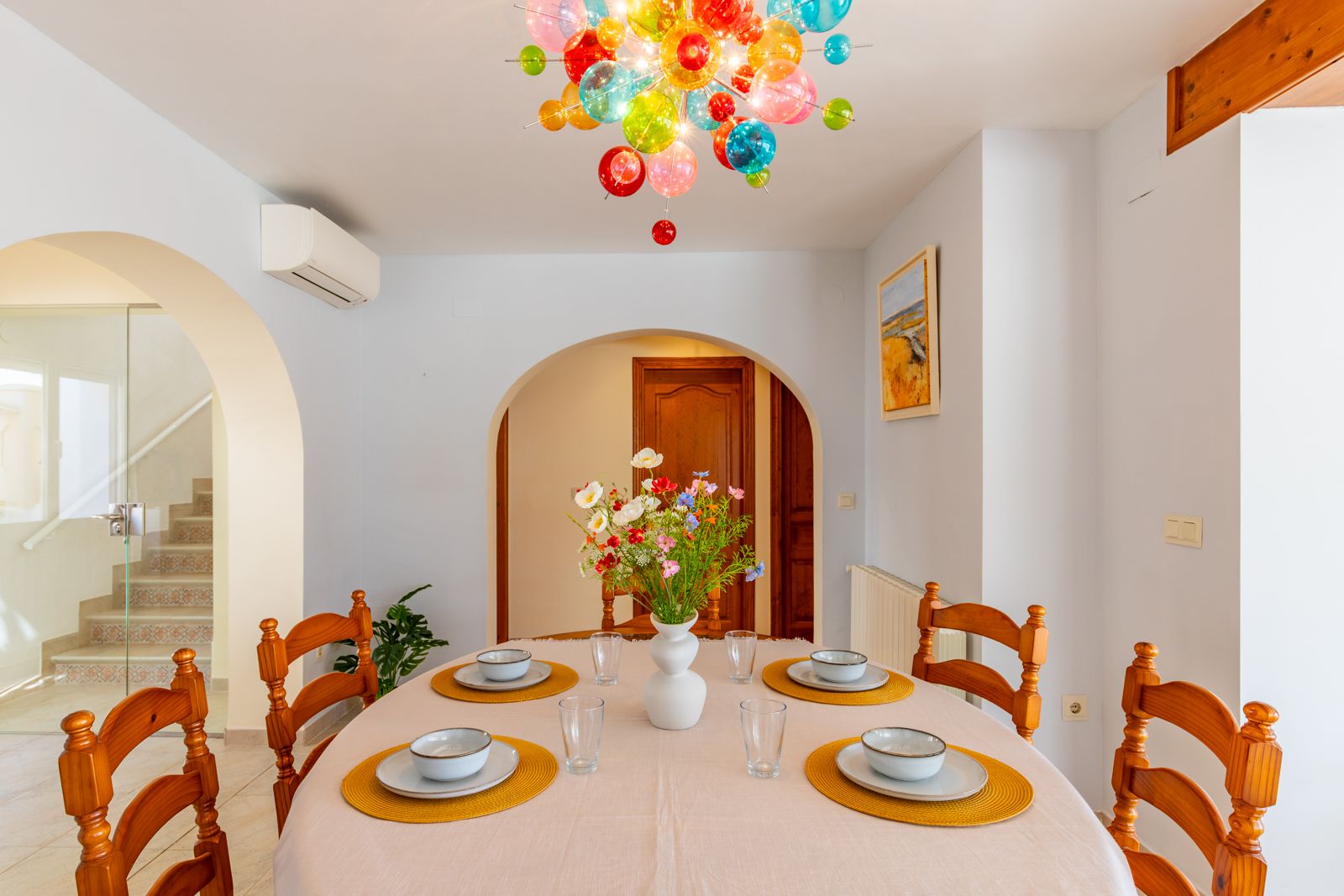 Salle à manger lumineuse avec table ovale et chaises en bois, ornée d'un lustre en bulles de verre colorées. L'espace se distingue par ses arches et son escalier aux carreaux décoratifs.