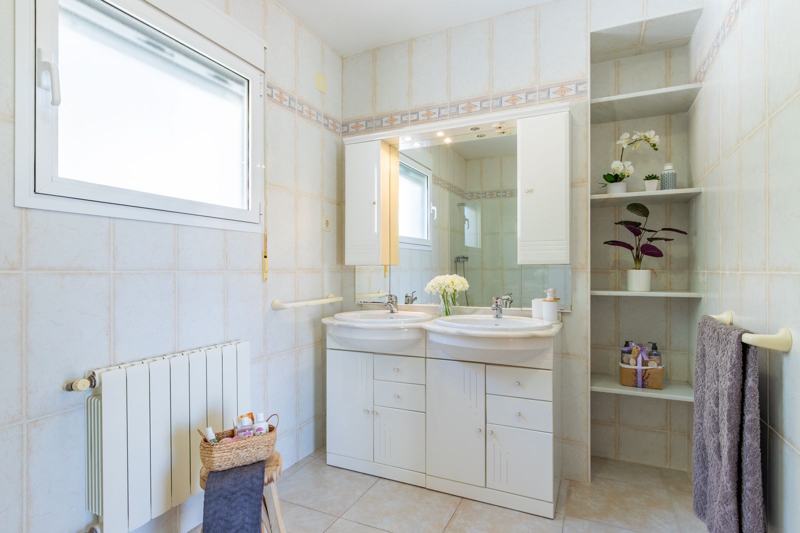 Salle de bain avec double vasque, meuble de rangement blanc, miroir éclairé et étagères encastrées. Murs carrelés avec frise décorative et radiateur mural.