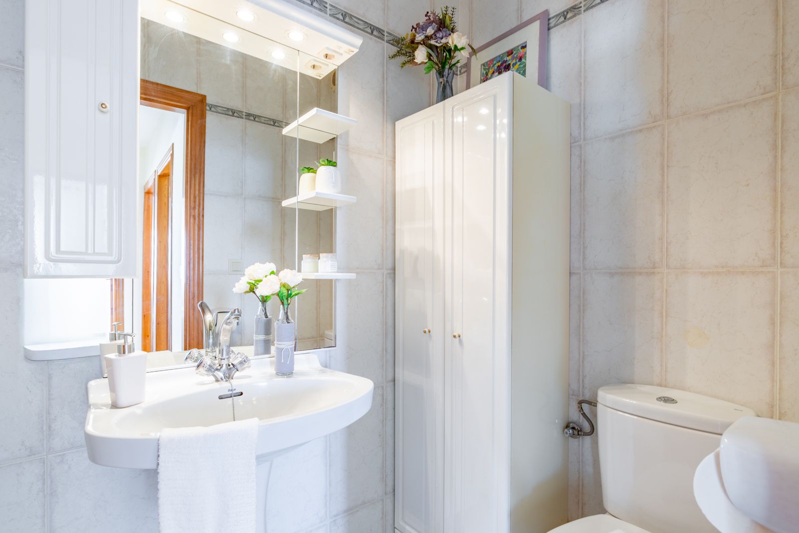 Salle de bain fonctionnelle avec lavabo sur colonne, miroir à éclairage intégré et étagères en verre, accompagnée d'un meuble de rangement blanc et de carrelage neutre.