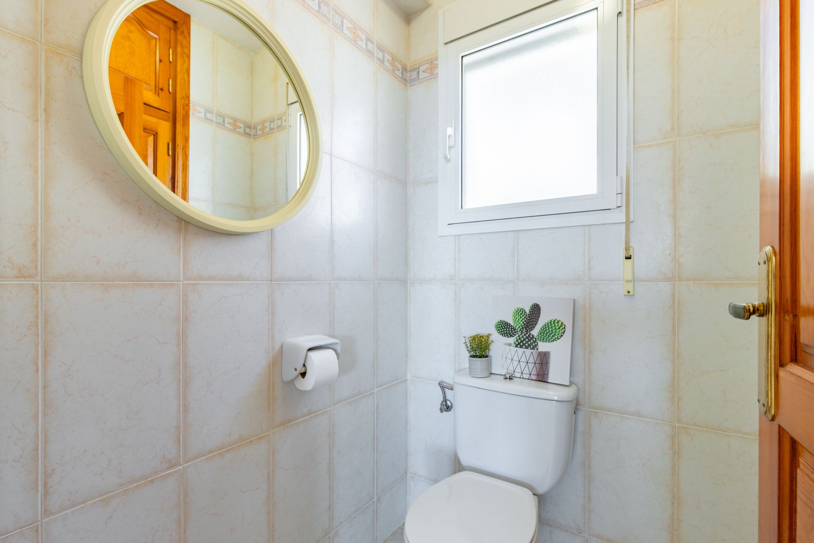 Toilettes fonctionnelles avec carrelage mural beige, miroir circulaire au cadre clair et fenêtre en verre dépoli offrant lumière naturelle et intimité.