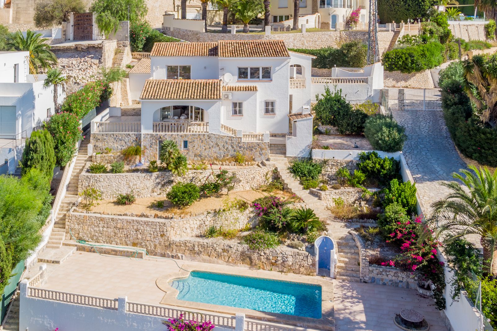 Vue extérieure de la villa avec façades blanches, toits en tuiles et jardins en terrasses avec murs en pierre naturelle menant à la piscine privée.