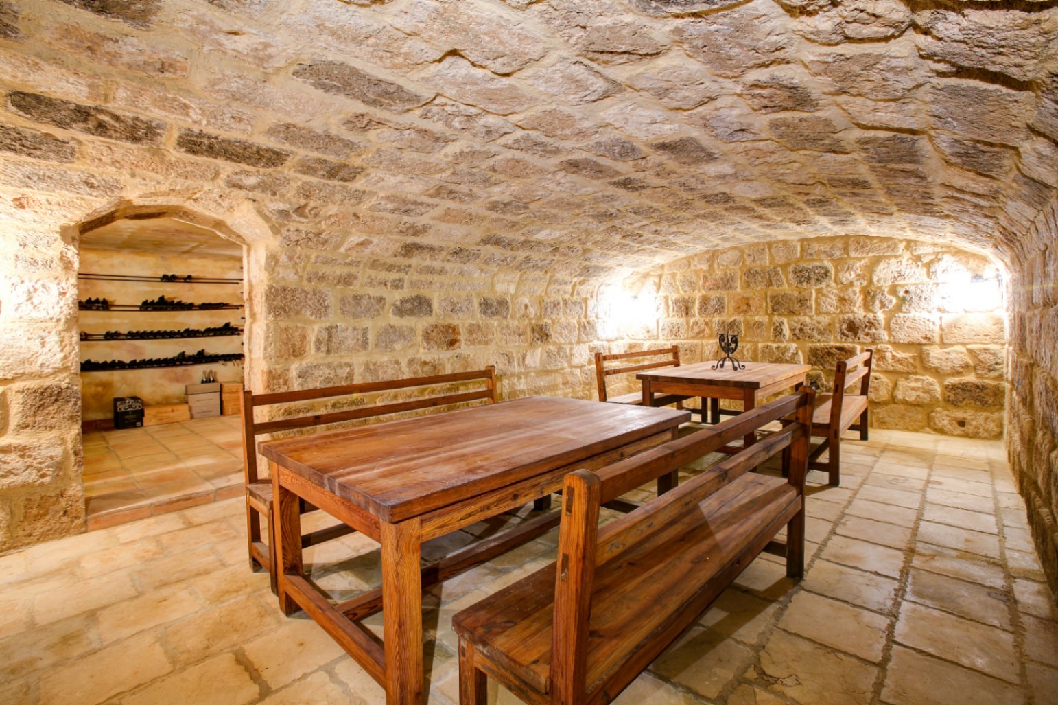 Cave à vin rustique avec plafond voûté en pierre, sol en terre cuite et tables en bois massif, complétée par un espace de stockage de bouteilles.