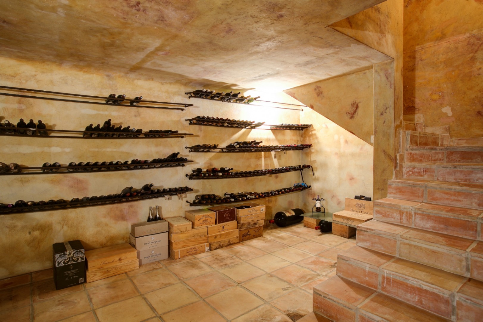 Cave à vin rustique avec sol en terre cuite, murs à la finition artisanale et casiers à bouteilles métalliques intégrés sous l'escalier.