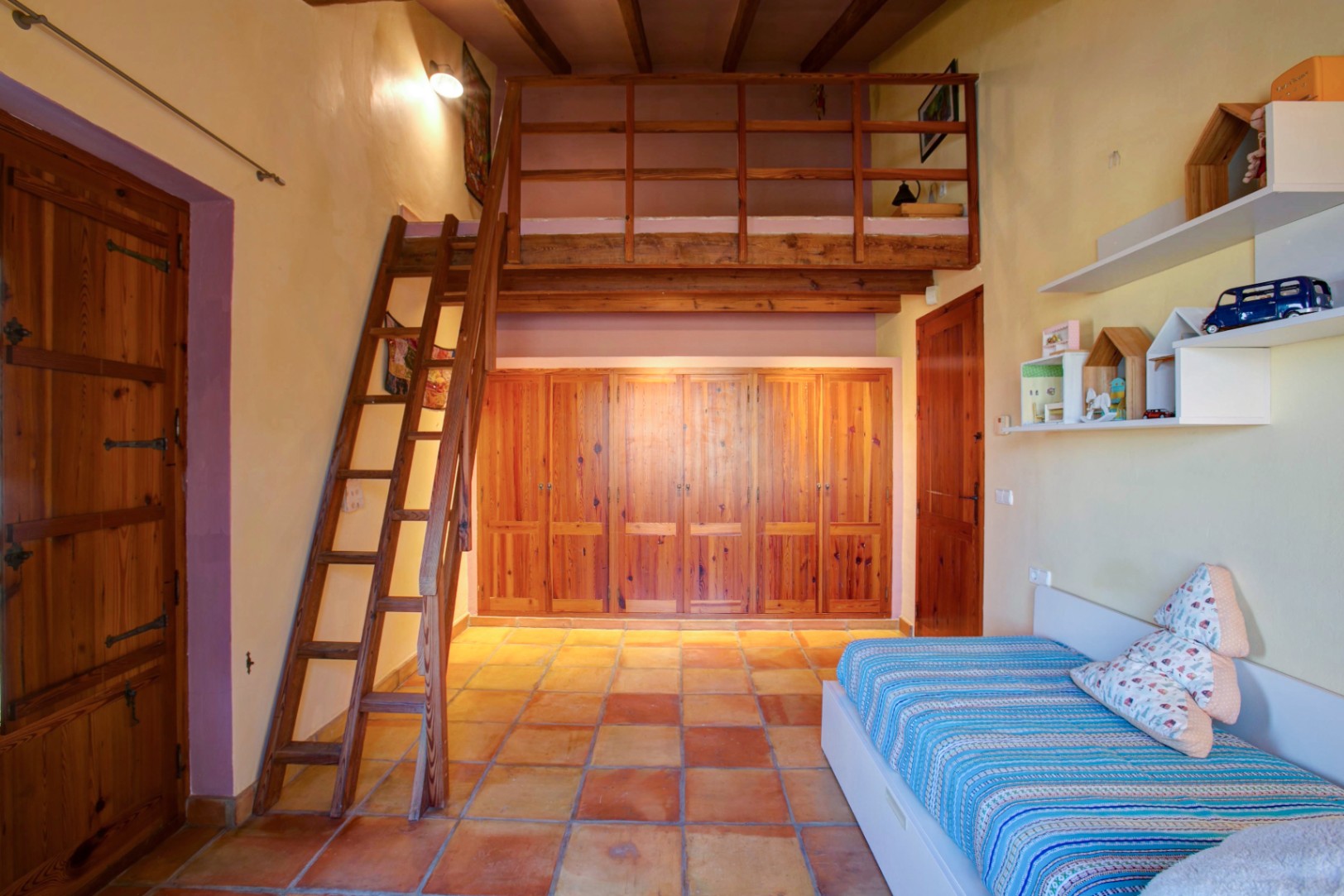 Chambre avec mezzanine en bois et échelle, placards intégrés en bois de mobila et sol en terre cuite traditionnelle sous poutres apparentes.