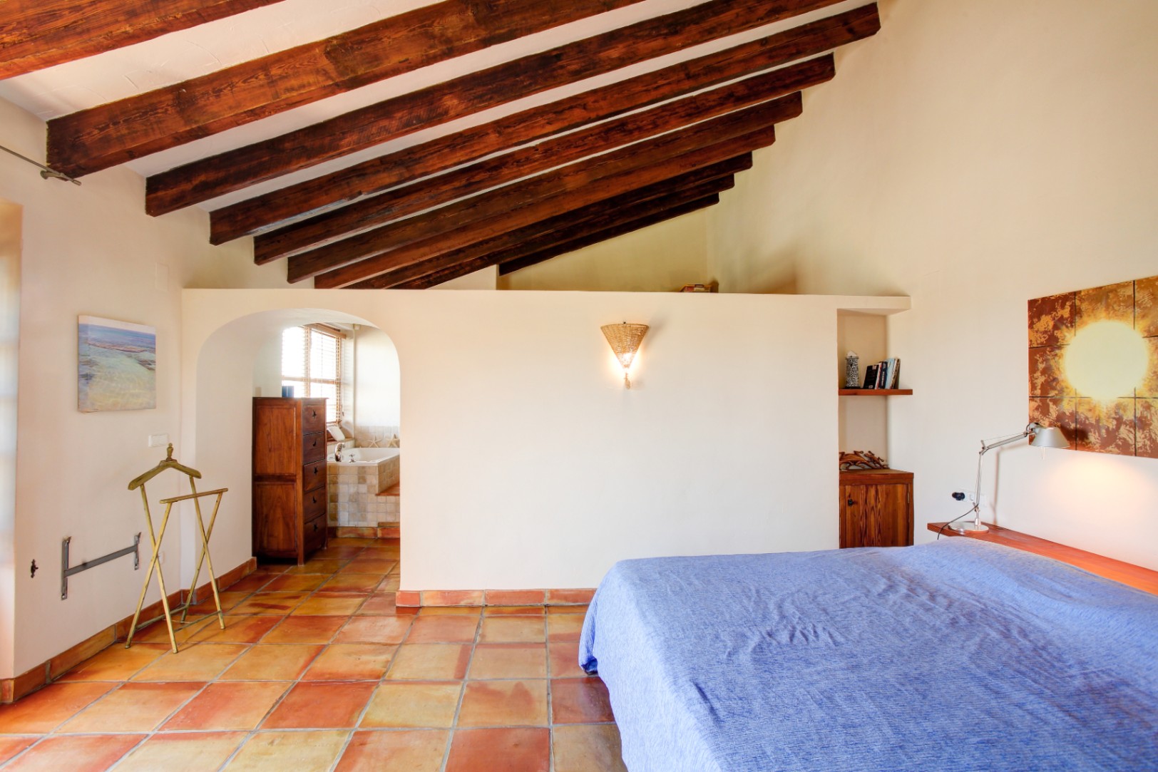 Chambre principale avec hauts plafonds aux poutres apparentes, sol en terre cuite et une arche élégante menant à la salle de bain attenante.