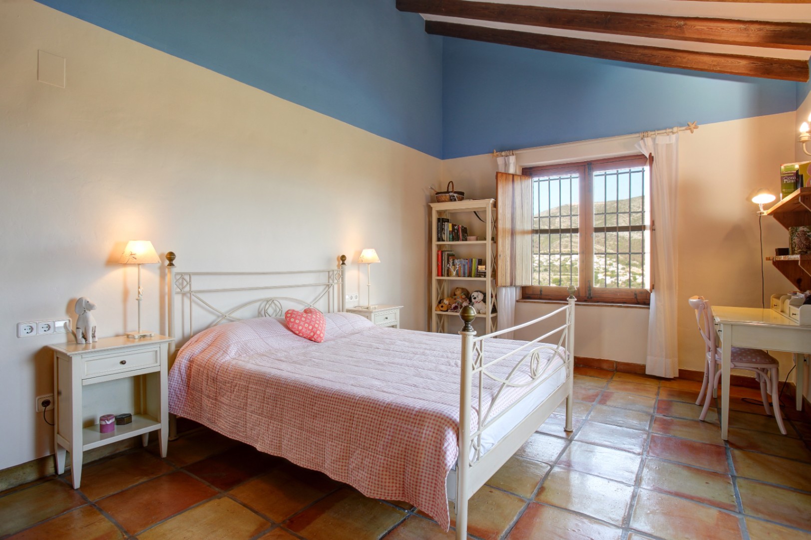 Chambre rustique avec poutres en bois apparentes, sol en terre cuite et lit en fer forgé blanc. Elle dispose d'un coin bureau et d'une vue dégagée sur les collines.