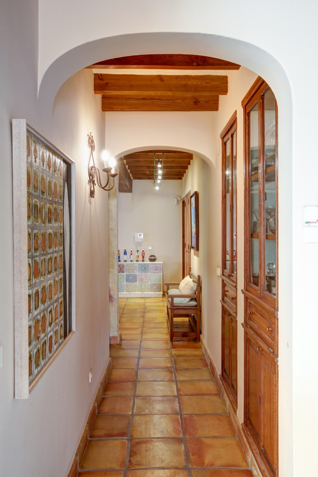 Couloir avec arches, poutres en bois apparentes et sol en terre cuite. Comprend des vitrines encastrées en bois noble et un éclairage mural de style rustique.
