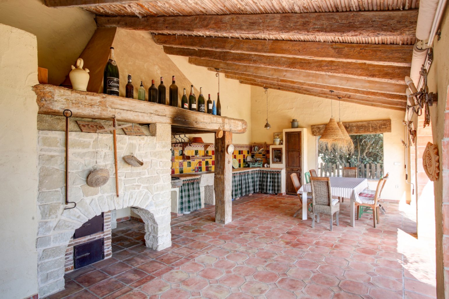 Cuisine d'été avec barbecue en pierre, poutres en bois massif et sol en terre cuite. Espace repas extérieur avec luminaires en fibres naturelles.